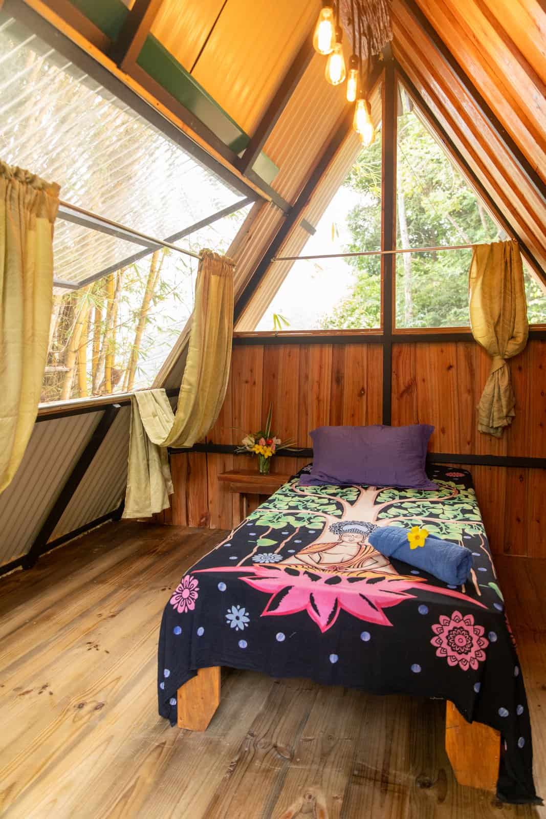 Inside an A-frame jungle cabin: warm hardwood walls, a bed dressed with a Buddha tapestry and triangular windows opening onto the canopy